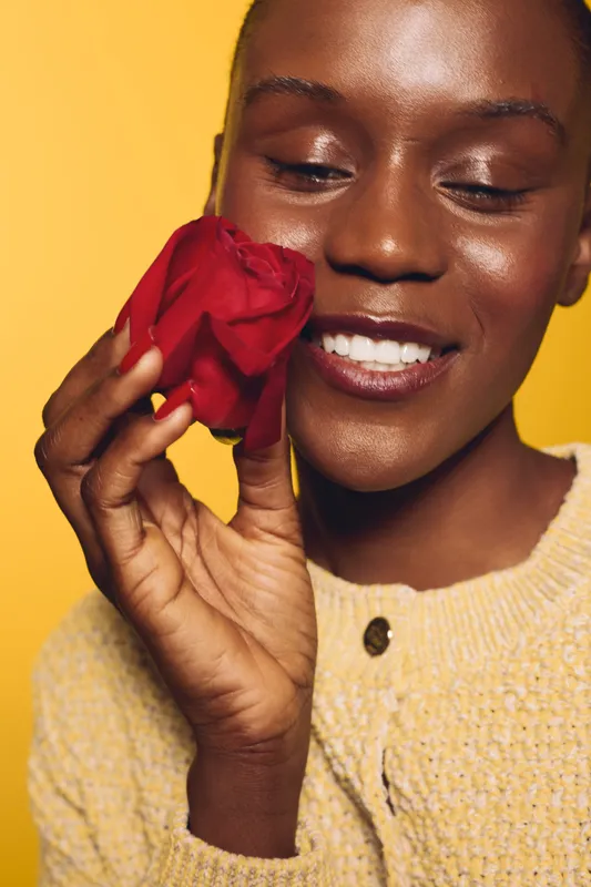 Close-up of a smiling dark skinned African woman holding a red rose near her face. She wears a yellow sweater against a yellow background. Concept of beauty, joy, and natural skincare.
