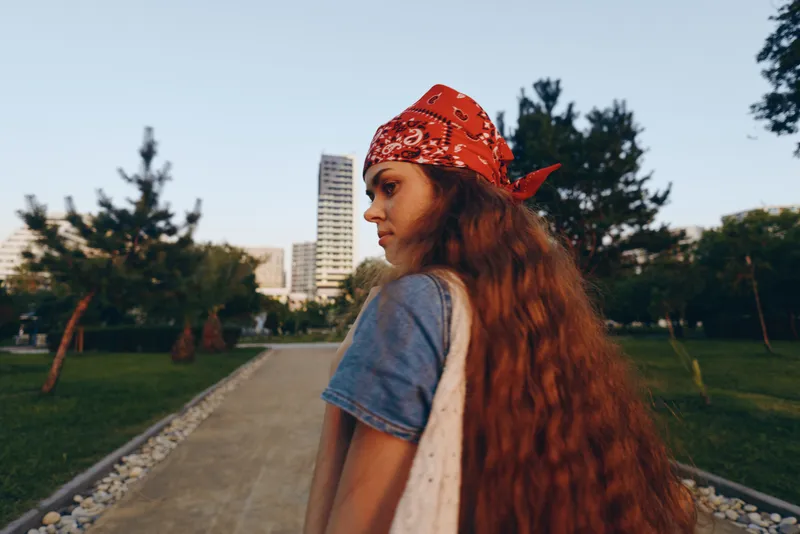 young woman with long hair and red bandana walking outdoors in casual clothes with city and park background during sunny day