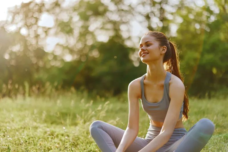 Young woman in sportswear enjoying nature, practicing yoga outdoors in a green park during sunny weather, embodying happiness and wellness