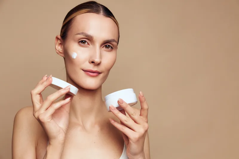 Young woman with clear skin applying white face cream on cheek. Close-up portrait on beige background. Concept of skincare, beauty, healthy lifestyle, self-care, wellness.