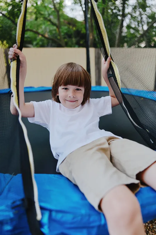 Happy boy enjoying his time on a trampoline, dressed in a casual white t-shirt and shorts, surrounded by greenery and vibrant summer vibes.