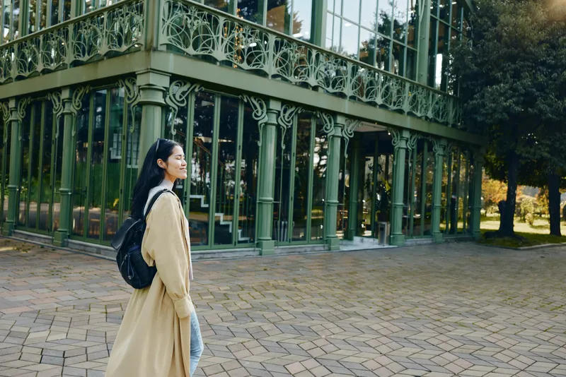 Woman with backpack in a beige coat strolling near a glass and iron greenhouse, urban park setting, soft light, candid style, travel mood, reflective moment, scenery calm atmosphere