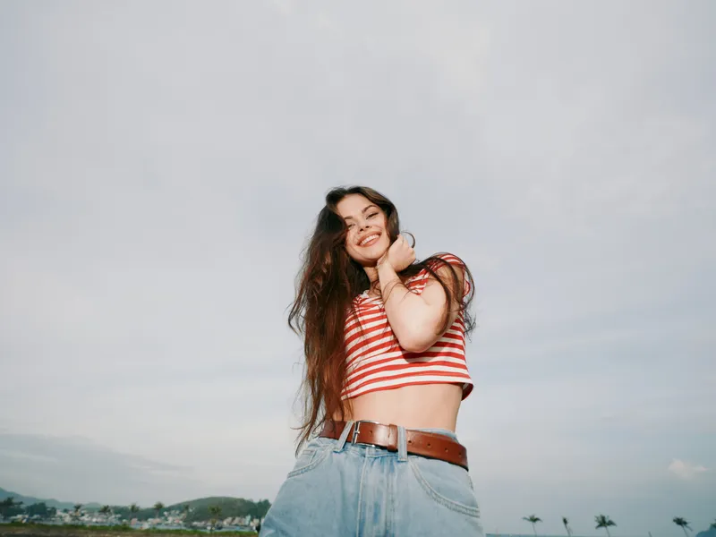 A joyful young woman stands outdoors under a bright sky, wearing a red and white striped crop top and high waisted jeans, smiling with a carefree pose.