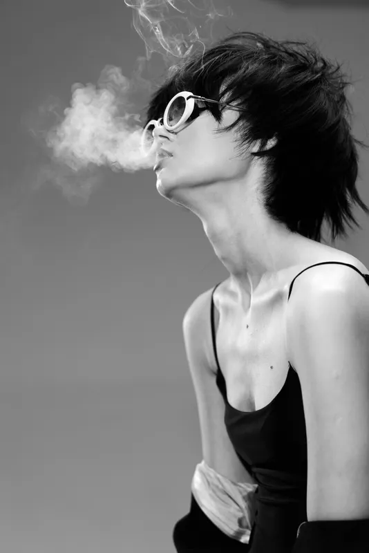 Black and white portrait of a stylish woman wearing sunglasses blowing smoke in a minimalistic setting.