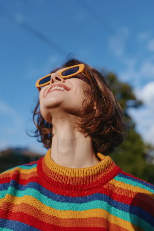 Young woman in a colorful striped sweater wearing orange sunglasses, smiling and looking up outdoors with short brown hair, joyful relaxed pose and sunny blue-sky lifestyle vibe.