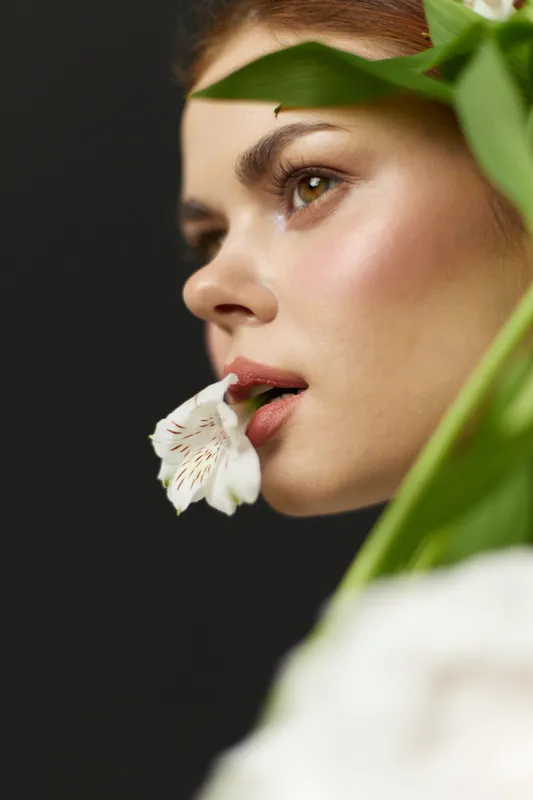 Young woman with natural makeup, holding a white flower, radiating serenity and elegance against a dark green backdrop, embodying beauty and tranquility