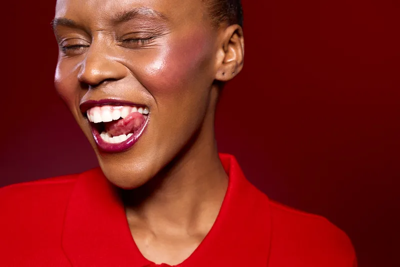 Close-up of a joyful black woman with dark skin wearing red clothing, eyes closed and tongue out, expressing happiness and playfulness on a red background. Concept of joy and energy.