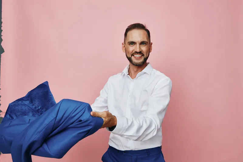 A professional man stands against a solid pink backdrop, holding a blue jacket. The isolated colored background highlights a confident, friendly business mood and a ready demeanor.