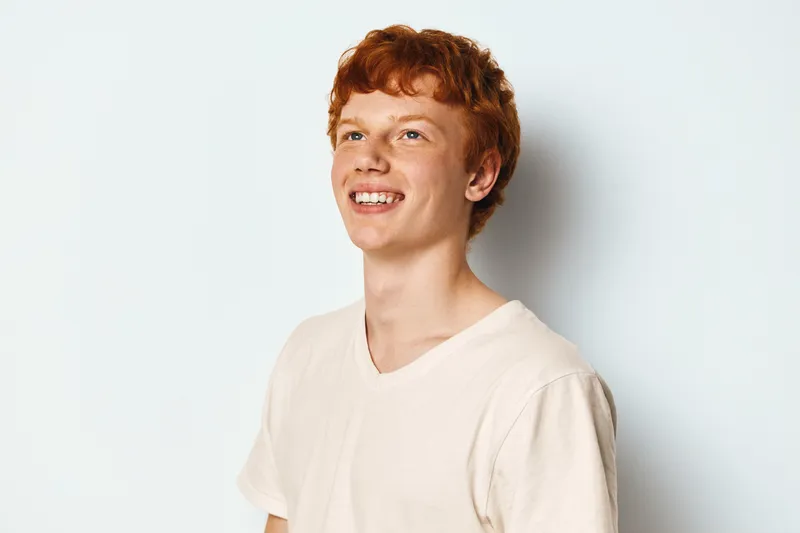 Young man with red hair smiling and looking up on white background with casual shirt. Portrait of happy male teenager with freckled face, natural expression and positive mood indoors.