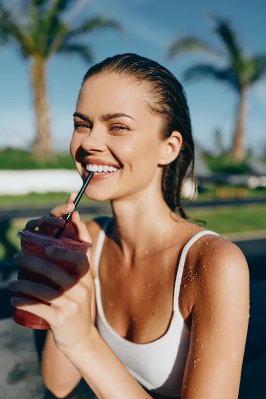 Young woman in a white tank top enjoys a smoothie outdoors on a sunny day near palm trees, smiling with a relaxed vibe as she sips through a straw