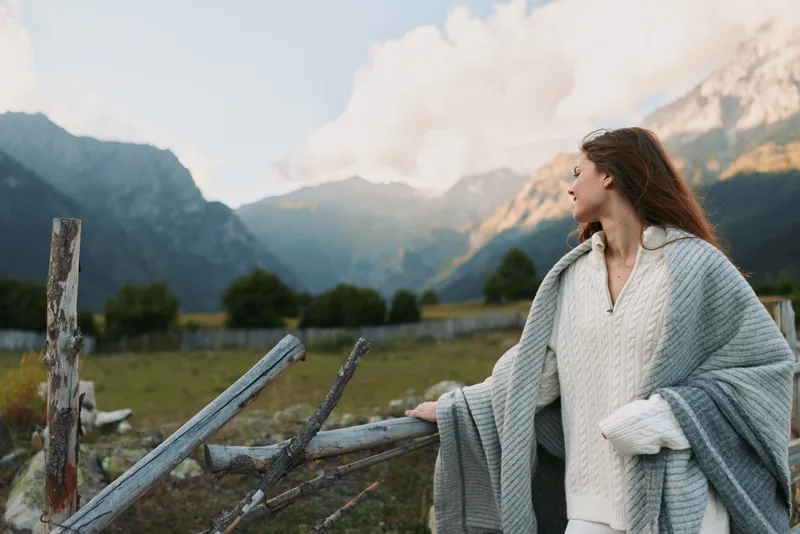 Young woman with long hair wrapped in a gray shawl stands by a rustic wooden fence looking at distant mountains under a partly cloudy sky during daytime.