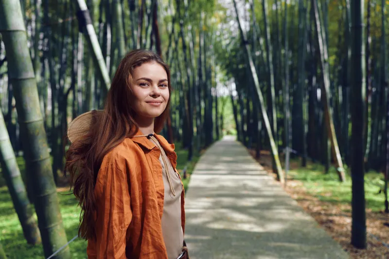 Woman bamboo forest path portrait nature outdoors smile, young female in orange jacket and hat standing on scenic walkway, longhair and sunlight among peaceful green foliage.