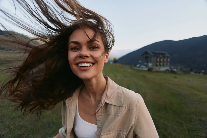 Young woman with windblown hair smiles at the camera in an open field with rolling hills, wearing a beige shirt over a white top, her relaxed pose and bright expression conveying carefree summer mood