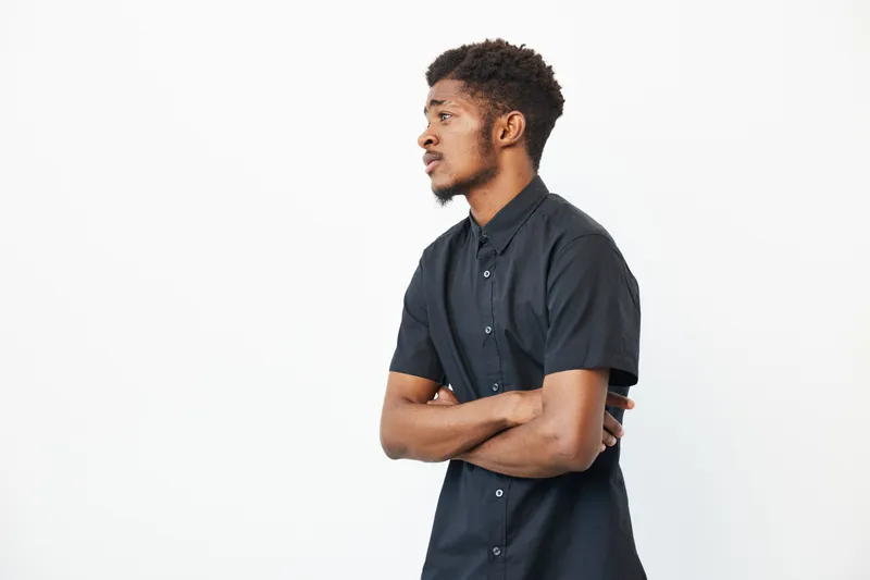 A thoughtful African American man stands confidently with arms crossed, showcasing a stylish black shirt against a clean, white background.