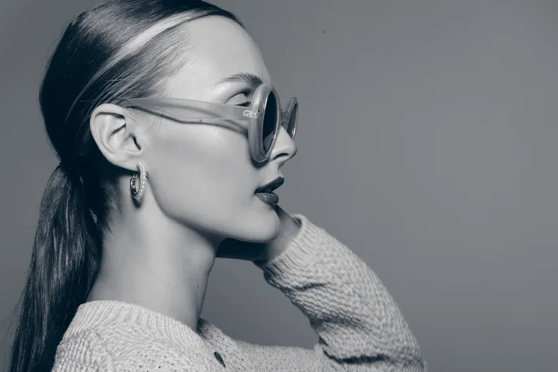 Profile of a young white woman with fair skin wearing sunglasses and a knitted sweater against a plain background.