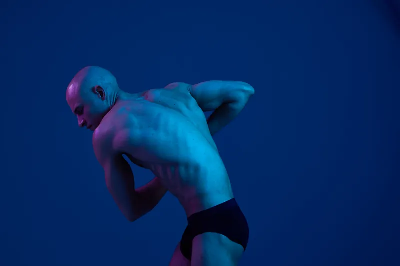 A strong man poses dramatically in a dark setting, showcasing his athletic physique against a moody blue background. The image has a modern, artistic feel.