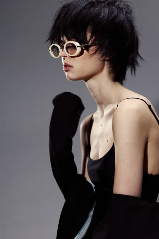 Stylish woman with short black hair wearing oversized sunglasses and a black outfit against a gray background, modern fashion concept.