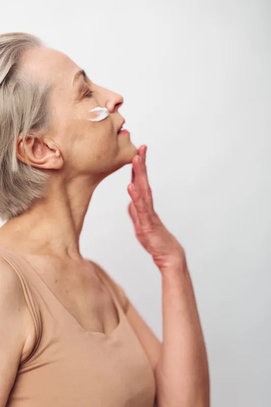 Senior woman with grey hair applies face cream to cheek, eyes closed in calm expression. Skincare, aging skin care, facial moisturizing, beauty routine, self-care concept.