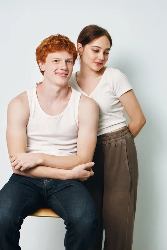Young couple posing together looking relaxed and comfortable in casual clothes against white background. Smiling man sitting on chair with woman standing close beside him.