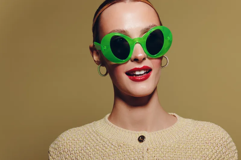 Stylish woman wearing large green sunglasses and red lipstick smiles confidently against beige background. Fashion portrait with trendy accessories and modern makeup concept.