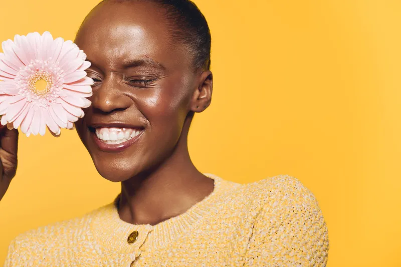 Happy African woman with dark skin smiles with closed eyes holding pink flower near face. Bright yellow background and cozy sweater create warm, joyful mood. Concept of beauty and nature.