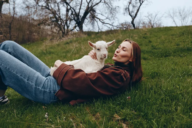 Young woman with brown hair lies on grass holding a white baby goat in her arms enjoying a peaceful moment outdoors in casual clothes during early spring.