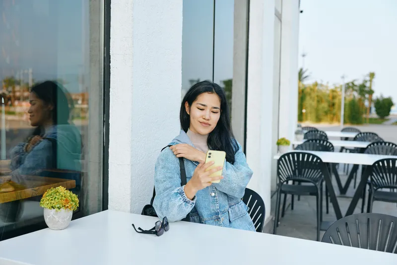 Woman sitting at a cafe terrace, checking a smartphone, sunglasses on the table, wearing a denim jacket, on a sunny day with a potted plant nearby, a relaxed urban moment of connection and casual