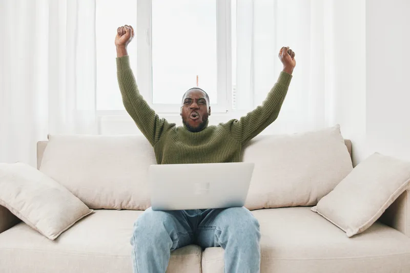 Happy Black man celebrating success while working on a laptop in a bright living room, wearing a green sweater with joy and excitement