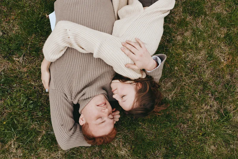 Young couple lying on grass embraces tenderly while wearing cozy sweaters enjoying a relaxed moment together in a natural outdoor setting with calm expressions.
