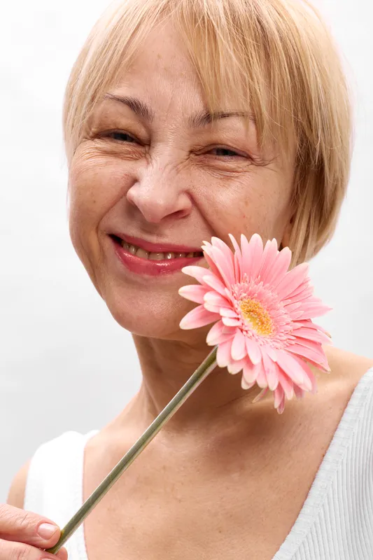 Woman portrait, flower smile, gerbera blonde closeup, spring mood, woman holding pink petals near face, soft white background, natural skin texture, gentle light, relaxed eyes