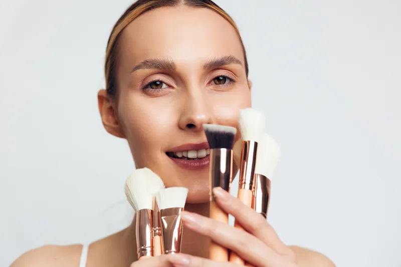 Woman applying makeup with a makeup brush, holding makeup brushes, close up portrait, beauty routine with cosmetics, soft studio lighting, white background, self care