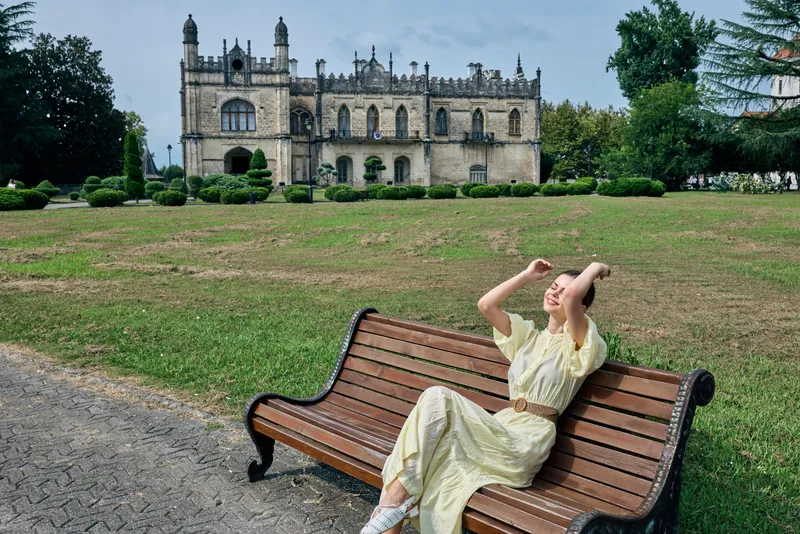 Young woman relaxing on a wooden bench in a sunny park with a historic castle in the background, wearing a light dress, enjoying a leisurely moment outdoors
