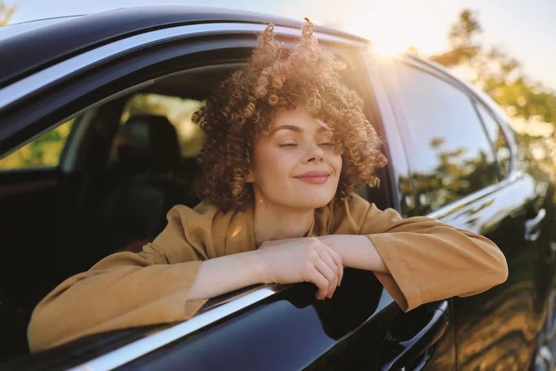 Young woman enjoys a calm drive, leaning on the car window with a soft smile, warm retro film tones, natural light, casual outfit, peaceful mood, outdoor setting, sun flare, reflective moment,