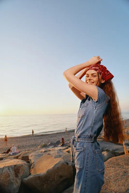 lifestyle woman in modern boho-western denim street style posing joyfully on rocky beach at sunset with film color tones creating warm, natural atmosphere