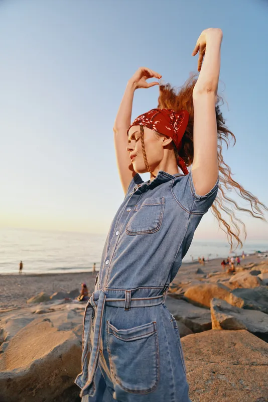 Lifestyle woman in modern boho-western denim street style, posing with a red bandana on beach during golden hour, film color tones enhancing natural beauty.
