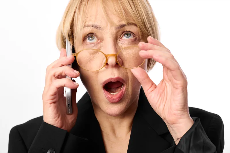 Surprised woman in business suit holds phone to ear and adjusts glasses on white studio background, showing stress, calling anxiety, face portrait closeup shot