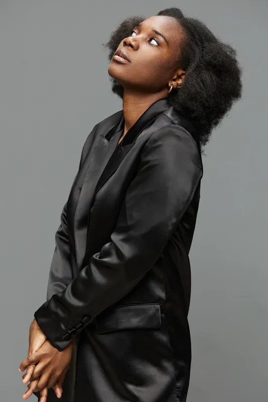 Young Black woman in a sleek black satin blazer, looking upwards with an expression of hope, set against a neutral grey background