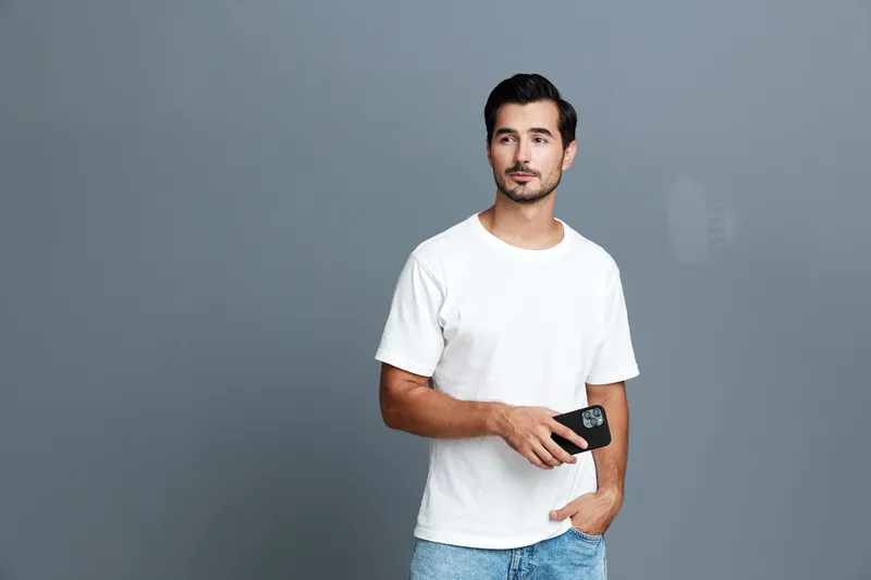Smiling man wearing white t-shirt holds smartphone in hand, looks at the camera with a confident expression, against solid gray background. Lifestyle concept.