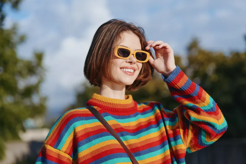 Woman smiling portrait outdoors in colorful striped sweater and yellow sunglasses with short brown hair, casual fashion and crossbody bag, sunny day, cheerful expression and street style vibe.