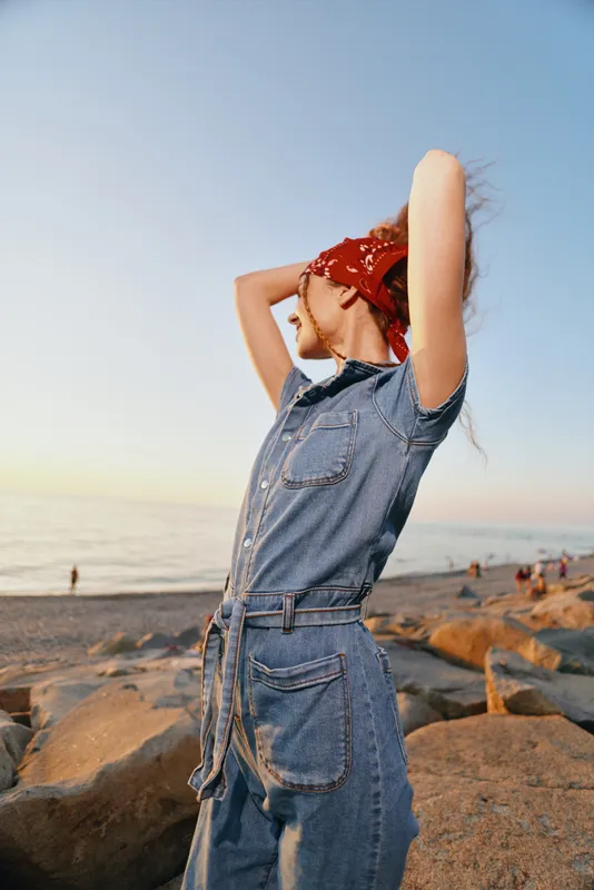 lifestyle woman in modern boho-western denim street style wearing a red bandana at the beach with warm film color tones and relaxed pose