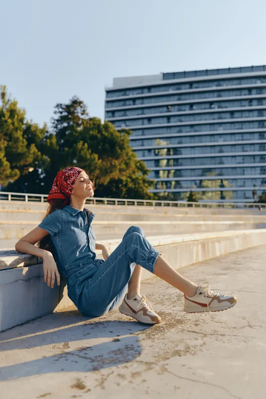Lifestyle woman in modern boho-western denim street style relaxing outdoors, wearing a red bandana and sneakers in bright natural light with film color tone.