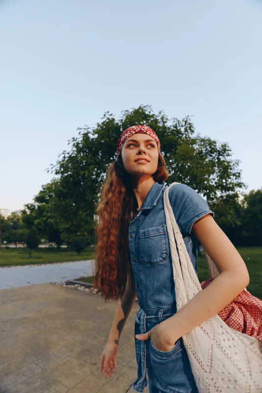 lifestyle woman with long hair in modern boho-western denim street style wearing red bandana and carrying crochet bag, film color outdoor park scene