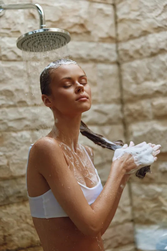 Young woman showering outdoors with closed eyes, washing wet hair with foam under rain shower, enjoying refreshing skincare routine in natural stone background.