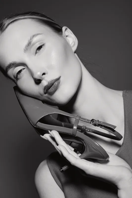 Caucasian woman with pale skin holds high heel shoe near face, posing in monochrome studio with soft lighting and elegant mood.