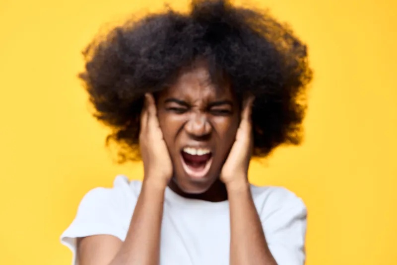 Young woman with curly afro hairstyle expressing frustration, holding her ears against a vibrant yellow background, showcasing emotions of distress and anxiety