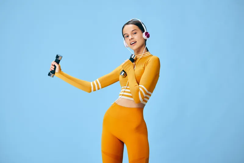 Young woman in orange fitness set lifts small dumbbells while wearing overear headphones, confident workout pose in a minimalist studio with solid pale blue background and soft lighting.