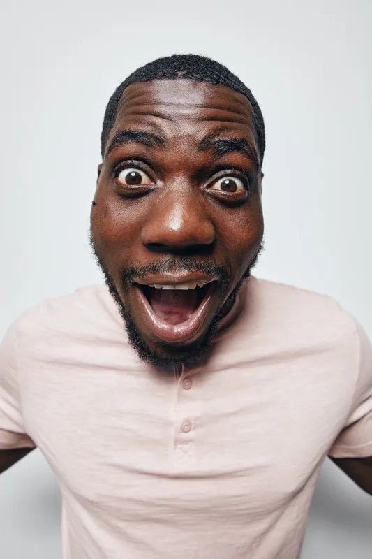 African man with dark skin and short hair wearing light pink shirt looking surprised with wide eyes and open mouth on white background studio portrait.