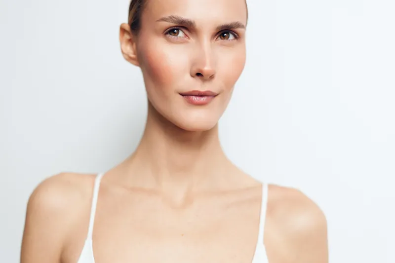 Closeup portrait of confident young woman with clear skin and natural makeup looking slightly away, wearing white top, minimalistic style, soft lighting, clean white background