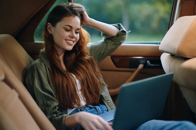 Young woman working on a laptop in a cozy car, showcasing relaxed emotions, casual clothing, and a serene outdoor background for remote work inspiration.