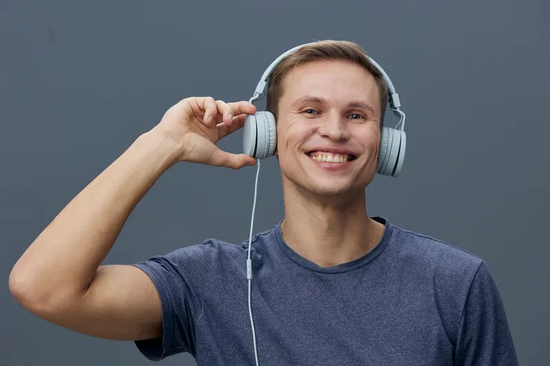 Happy young man with short brown hair wearing casual blue t-shirt listening to headphones smiling in front of gray background studio portrait. People lifestyle concept, entertainment, music, technolog
